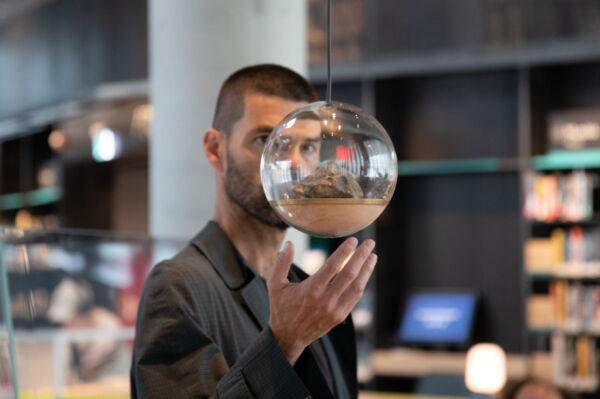 En person står bak en glasskule. Personen er inne på et bibliotek. Foto.