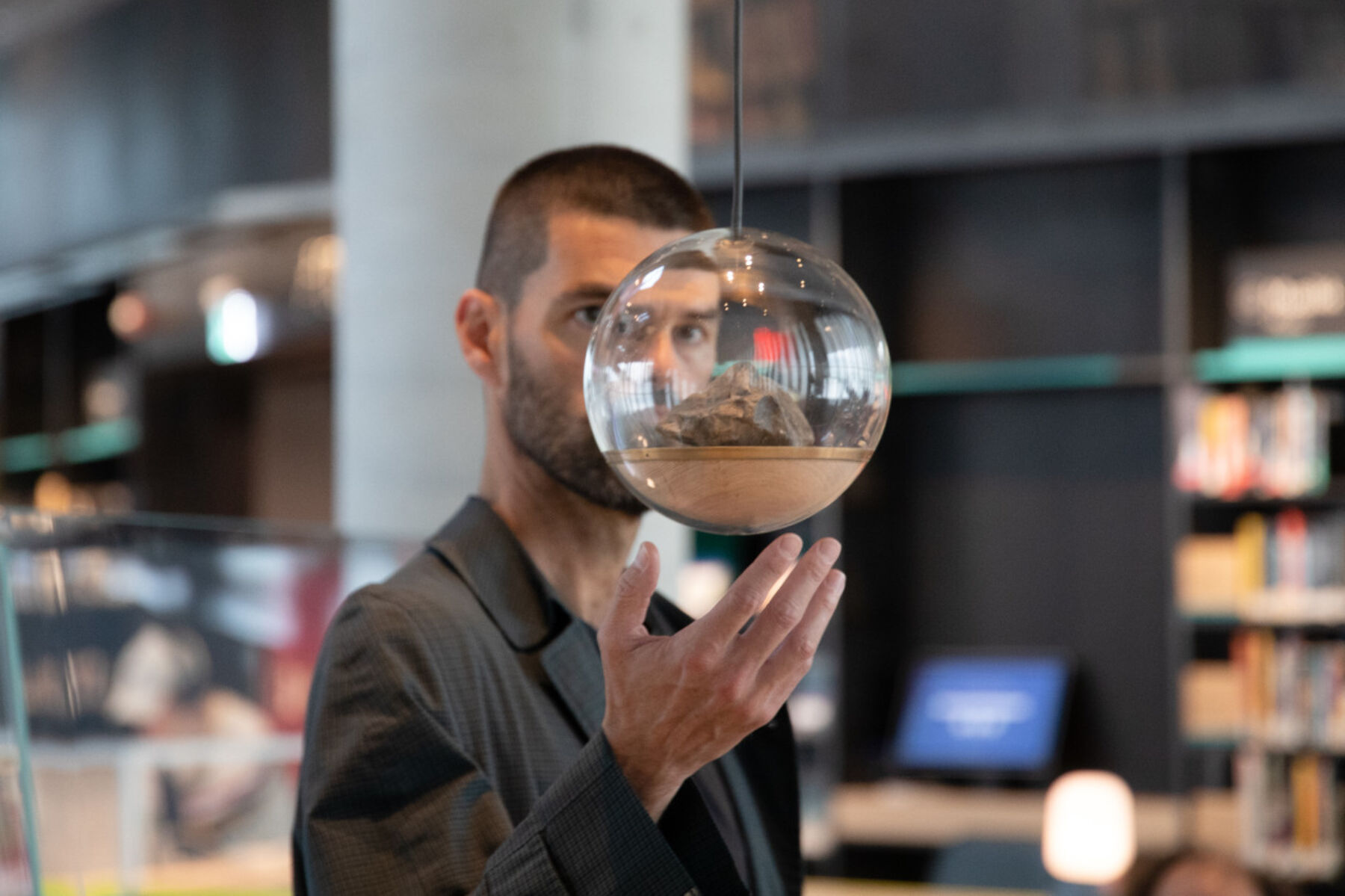 En person står bak en glasskule. Personen er inne på et bibliotek. Foto.