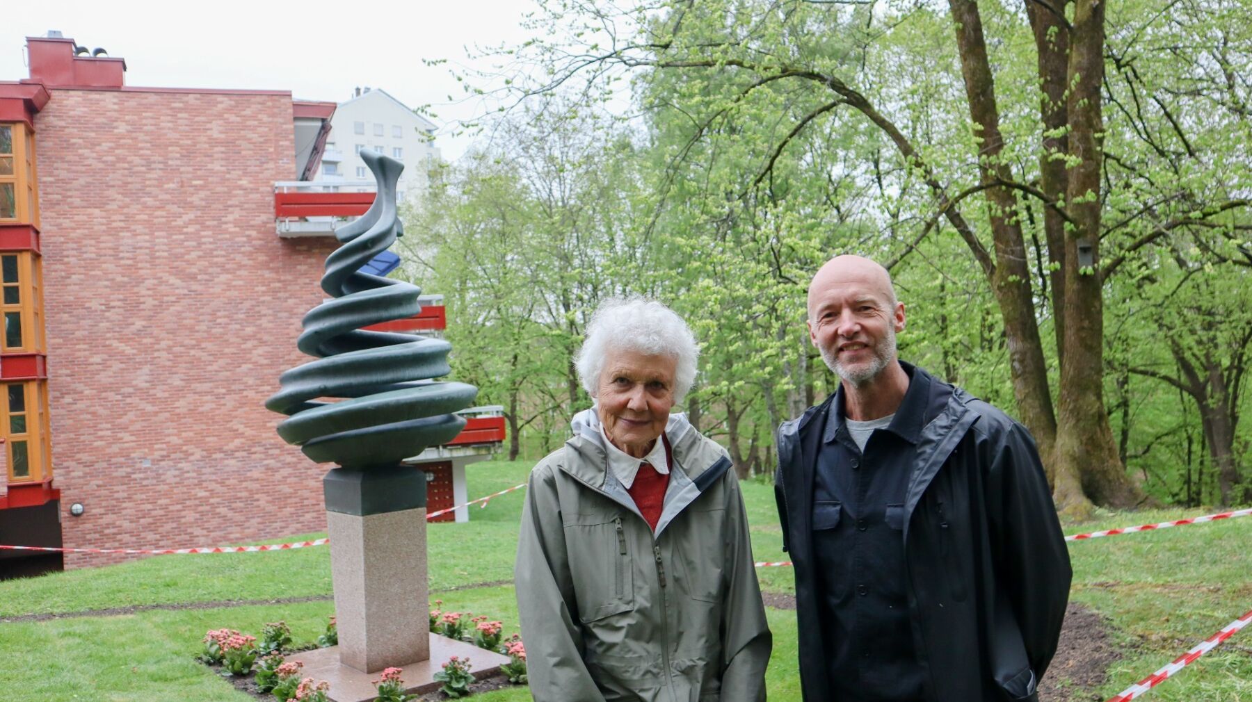 To personer står ved siden av hverandre. I bakgrunnen er en skulptur og en liten boligblokk.