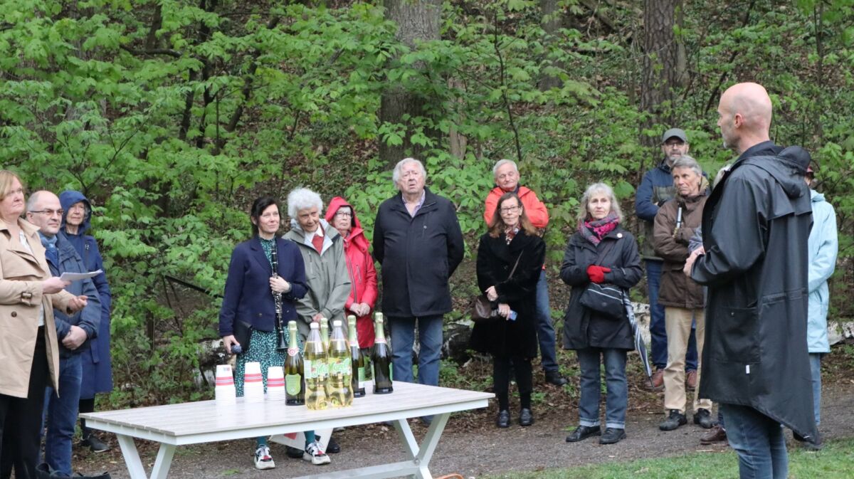 En liten mengde mennesker står og ser på en person som holder en tale. På bordet i mellom dem står flasker med drikke. Foto.