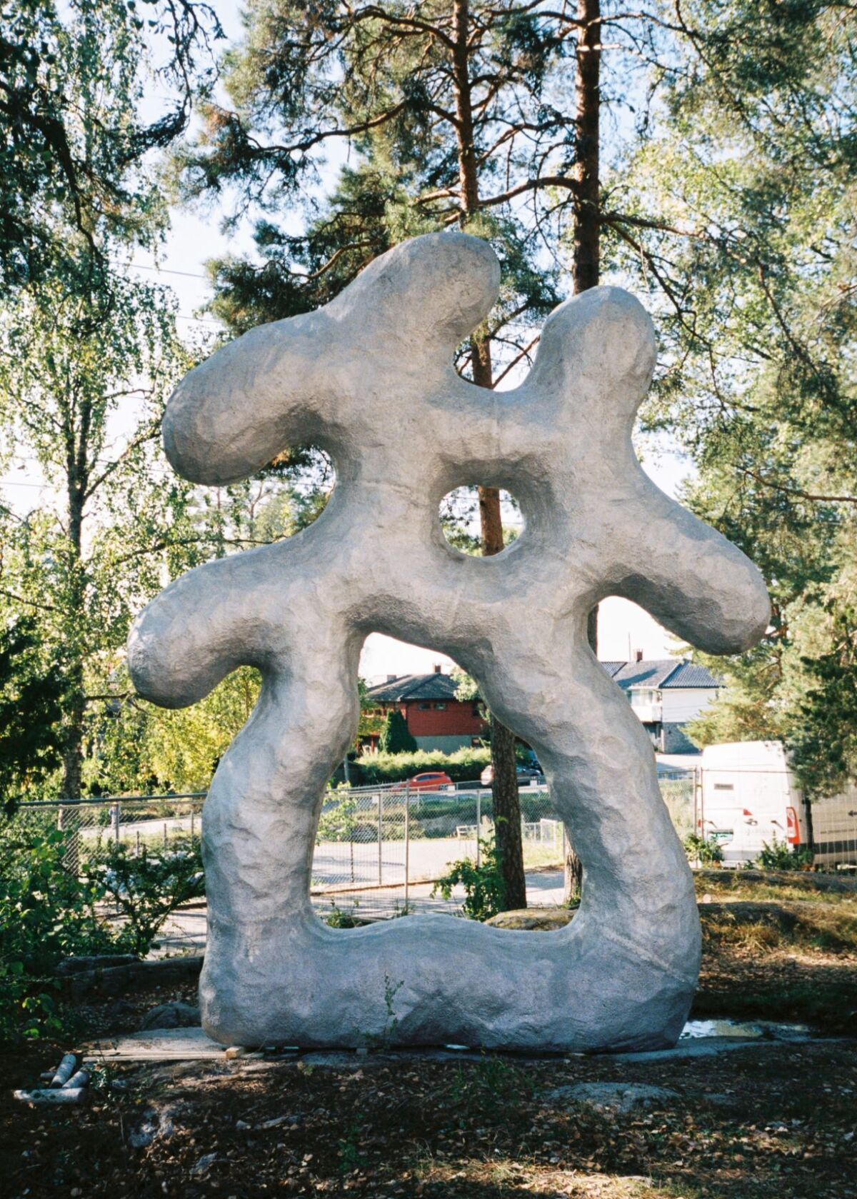 Lys grå skulptur står plassert i naturen med trær rundt. Foto.