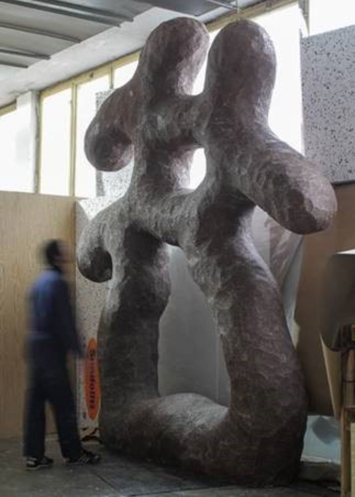 En person står foran og jobber med en stor skulptur innendørs i et verksted. Foto.