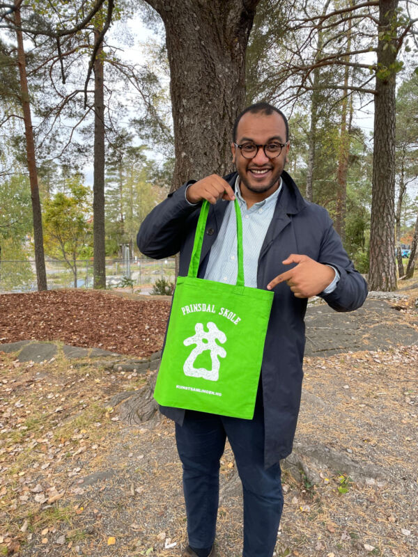 En person står utendørs og holder i en grønn tøyveske. Personen viser vesken frem med en hånd, og peker på den med den andre. Foto.