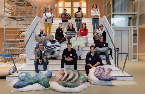 Fire personer står i en trapp. Foran dem sitter elleve personer. Personene holder i ulike organiske former og figurer.