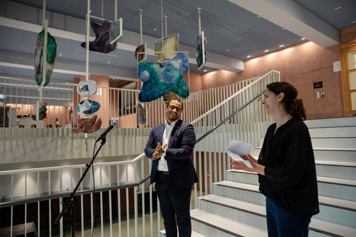 To personer står foran et kunstverk bestående av organiske former som henger fra taket. De klapper med hendene. Foto.