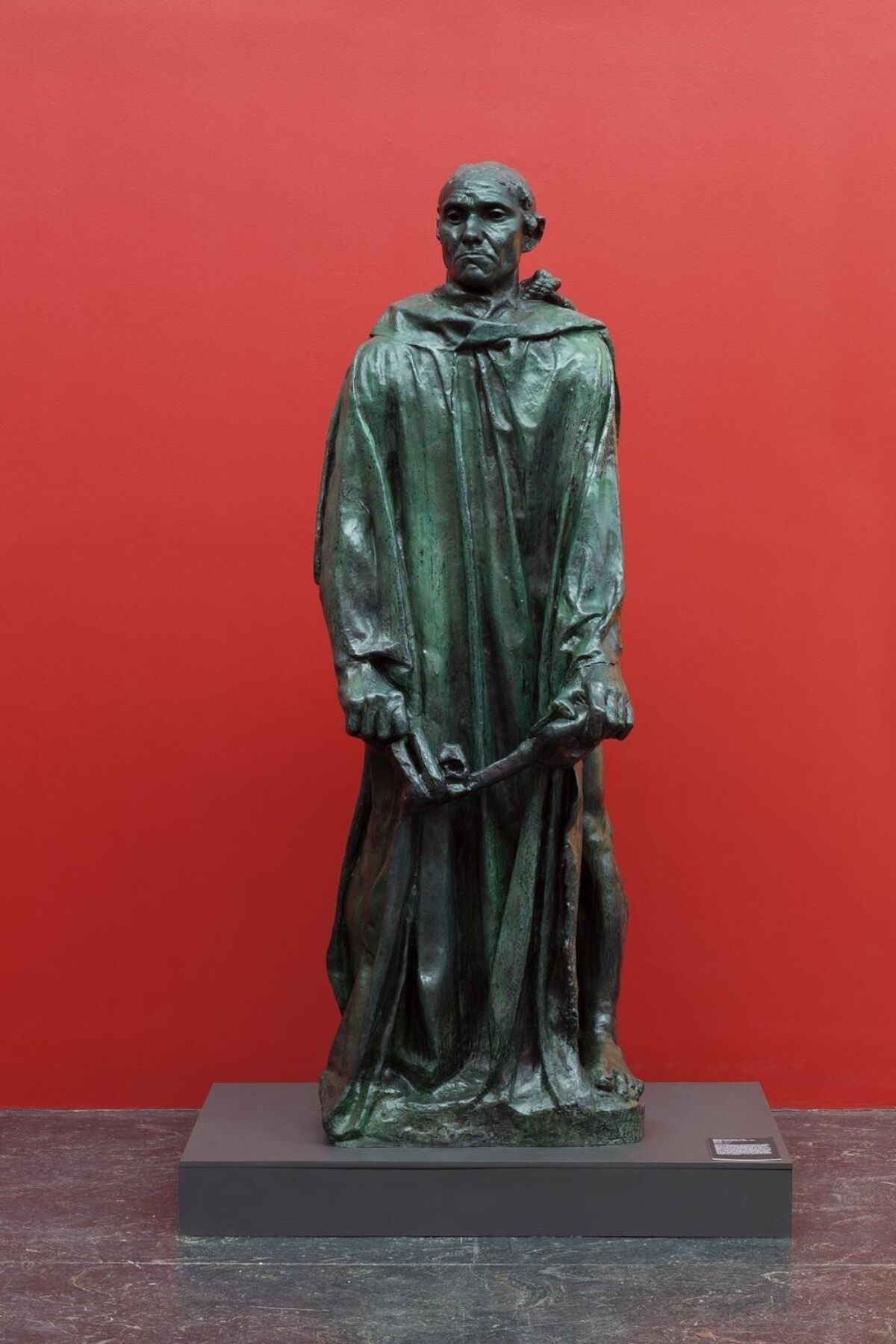 Skulptur av mann som står foran en rød vegg. Foto.