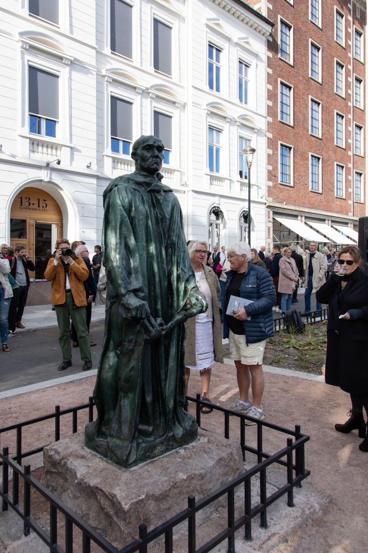 En skulptur av en mann står i midten av bildet. Rundt står mennesker og ser på. Foto.