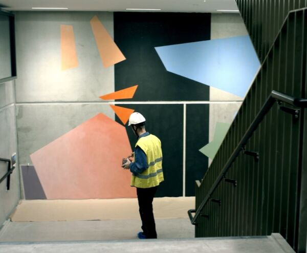 En person med hjelm og refleksvest står foran et fargerikt veggmaleri i en trappeoppgang. Foto.