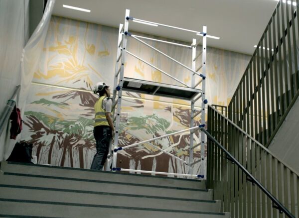 En person står sammen med et stillas foran et veggmaleri i en trappeoppgang. Foto.