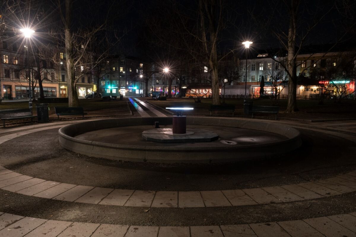 En parkplass utendørs med fontene og kunstverk som lyser i mørket. Foto.