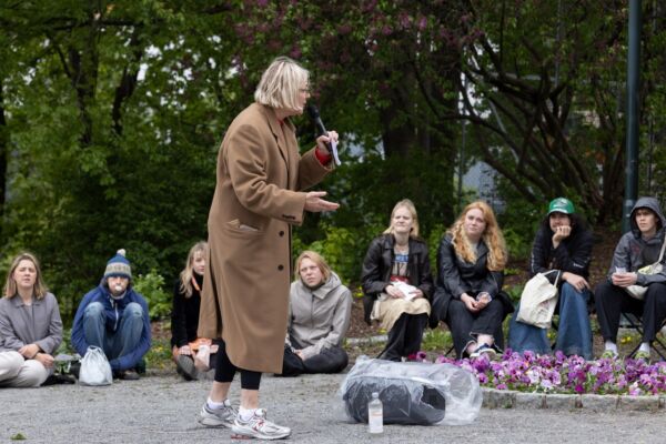 En person holder foredrag foran menneskemengde som sitter rundt. Foto.