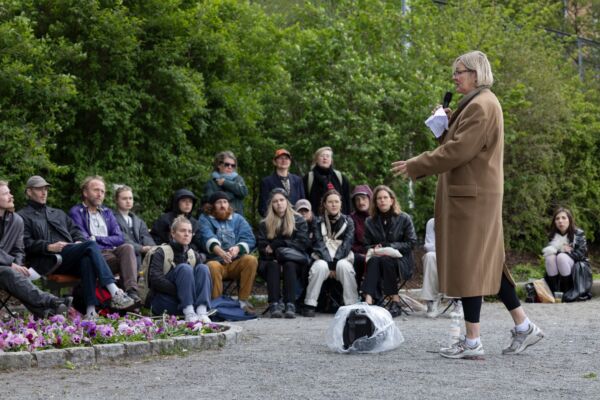 En person holder foredrag foran menneskemengde som sitter rundt. Foto.