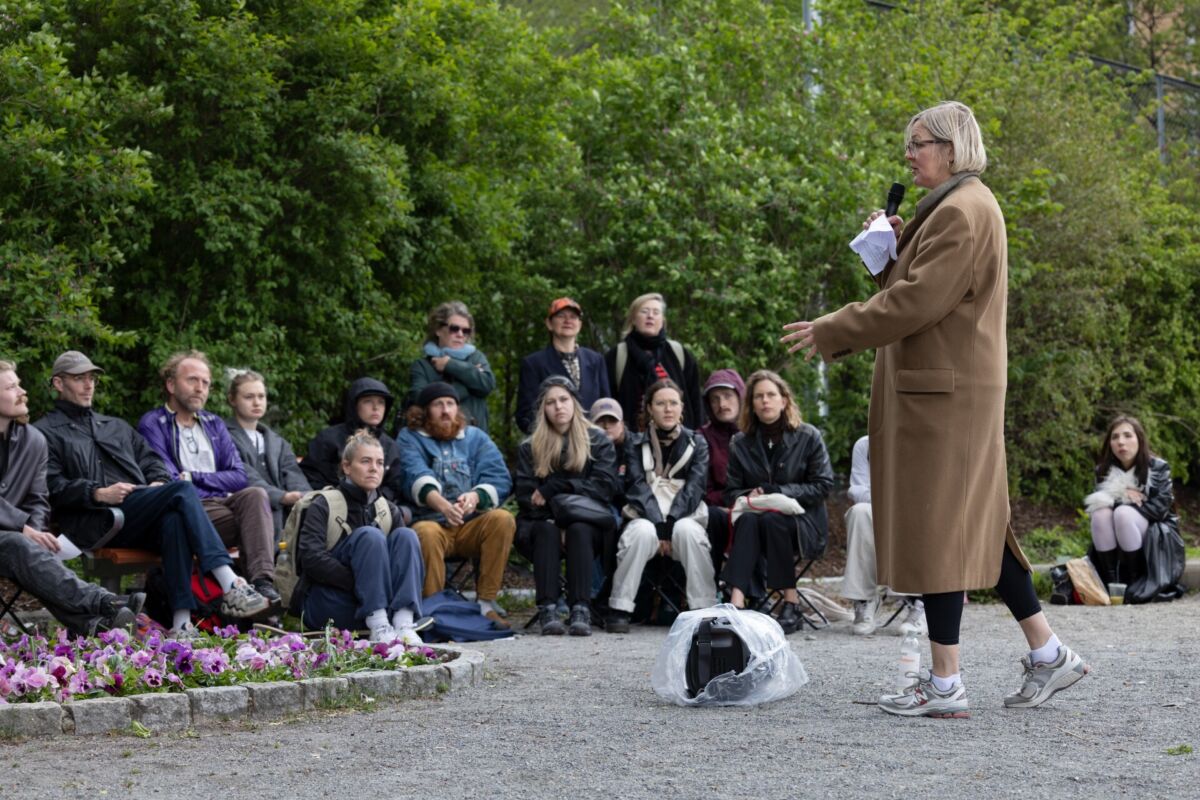 En person holder foredrag foran menneskemengde som sitter rundt. Foto.