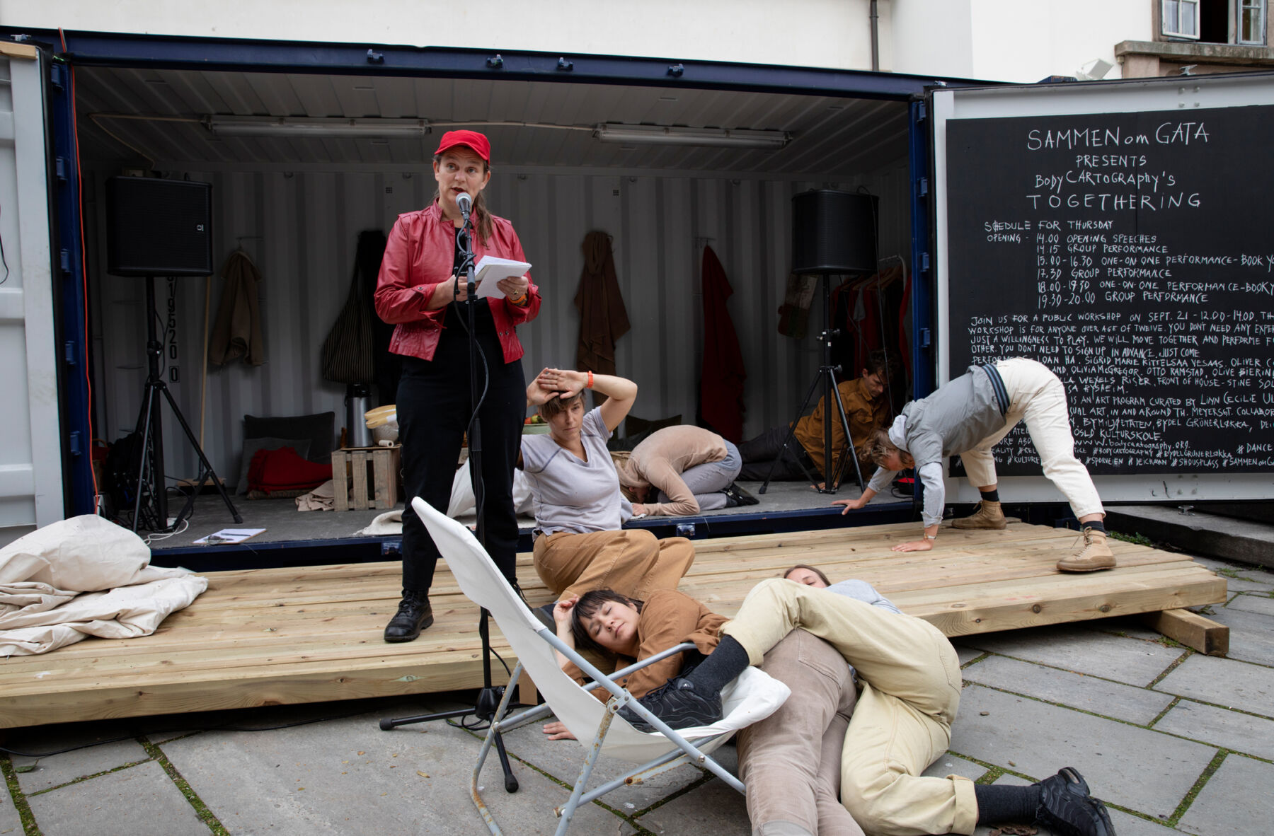 En person står og snakker i en mikrofon foran en container. Seks personer ligger, sitter eller krabber på bakken rundt. Foto.