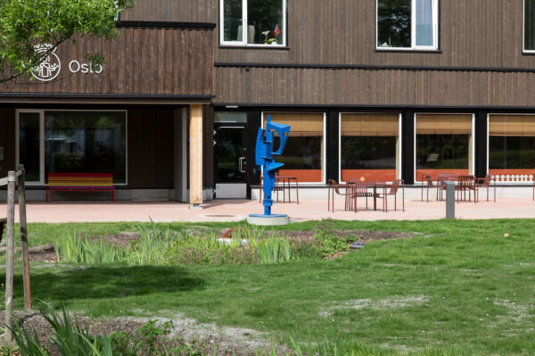 Grønt gress i forgrunn, brun bygning i bakgrunn, terasse med sittemøbler og en blå skulptur med abstrakt form.