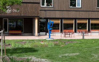 Grønt gress i forgrunn, brun bygning i bakgrunn, terasse med sittemøbler og en blå skulptur med abstrakt form.