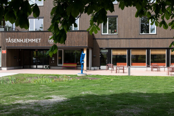Grønt gress i forgrunnen, brun trebygning i bakgrunnen med påskrift Tåsenhjemmet, bord, stoler og en blå skulptur ved inngangspartiet. Foto
