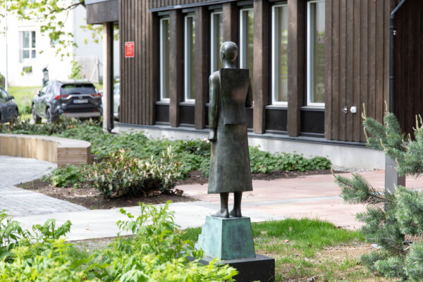 Skulptur av person i bronse, står på sokkel i uteområde foran brun bygning. Skulpturen er laget av Istvan Lisztes. Foto.