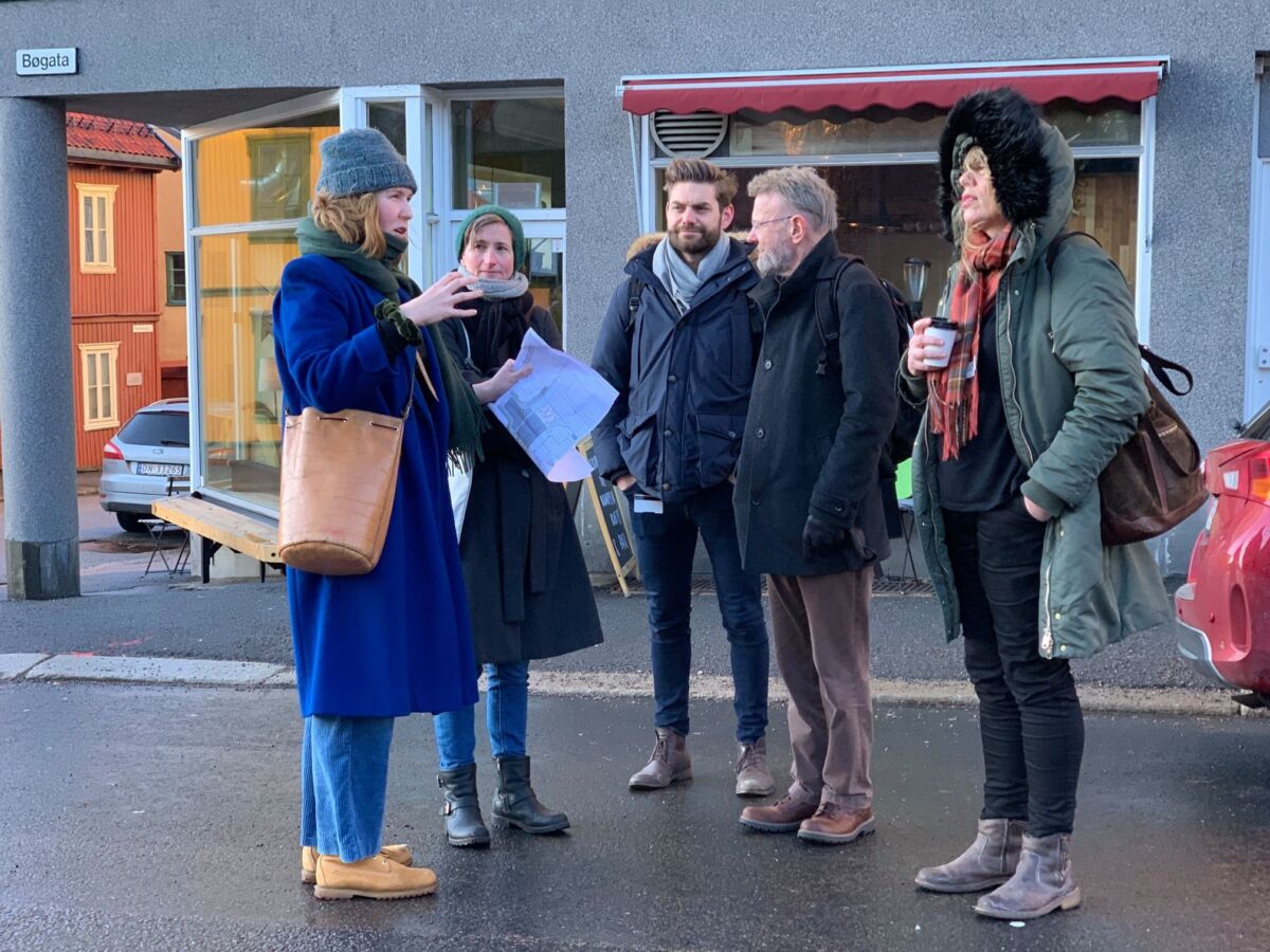 Fem personer står sammen og prater og ser på hverandre foran en kafébygning. Foto.