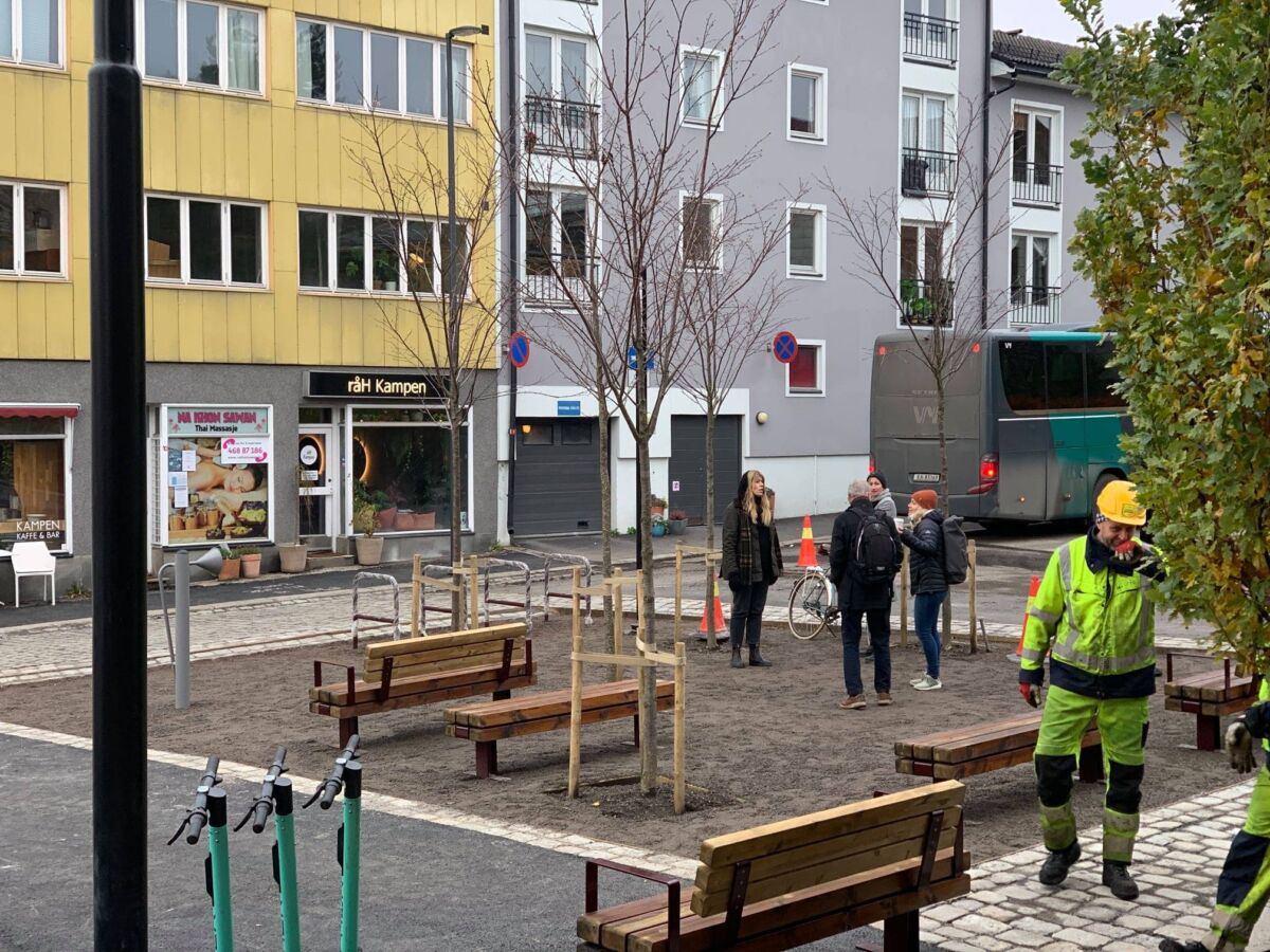 Et kirketorg med bygninger rundt, benker og sparkesykler. To personer i arbeidstøy står sammen på høyre side, og i midten står tre personer samlet. Foto.