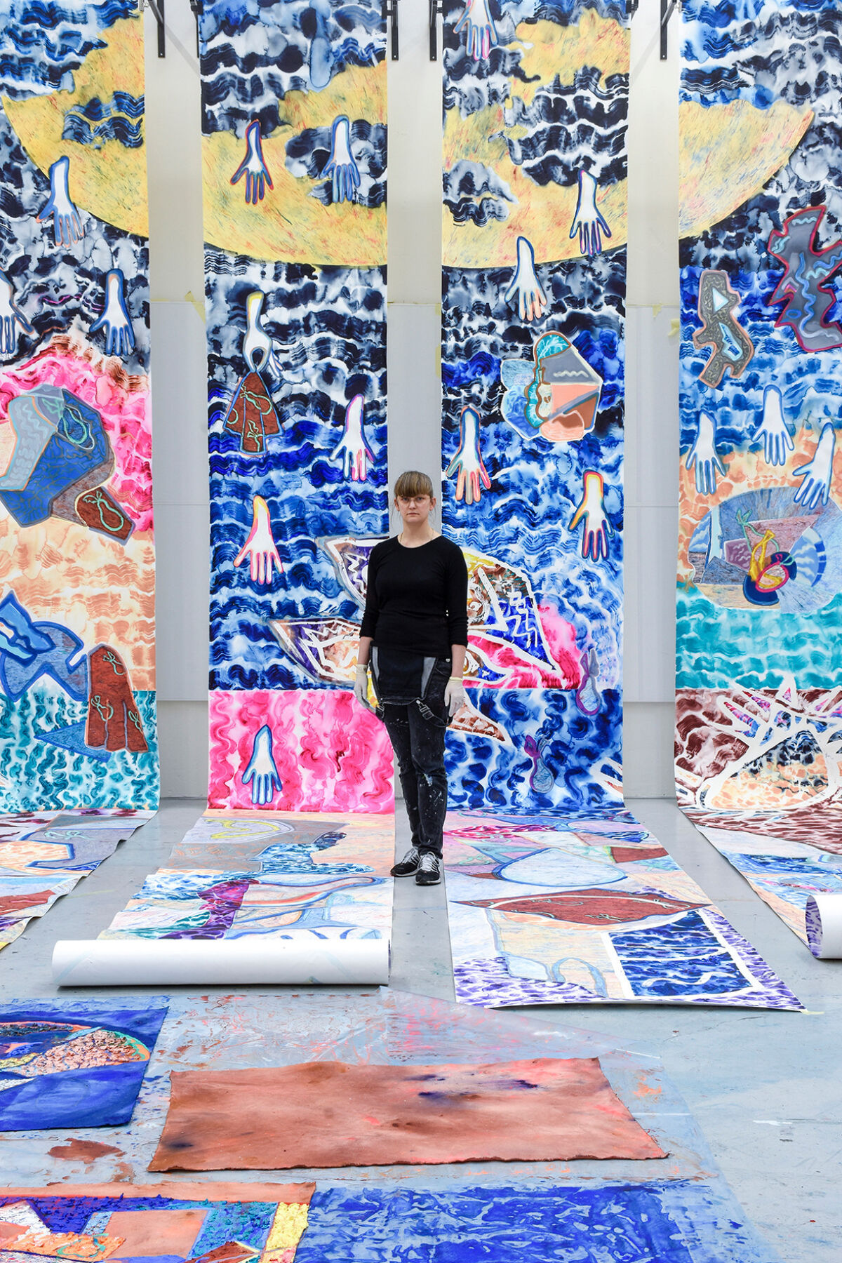 En person står i et rom omgitt av farge- og detaljrike kunstverk, både hengende i taket og liggende på bakken. Foto.